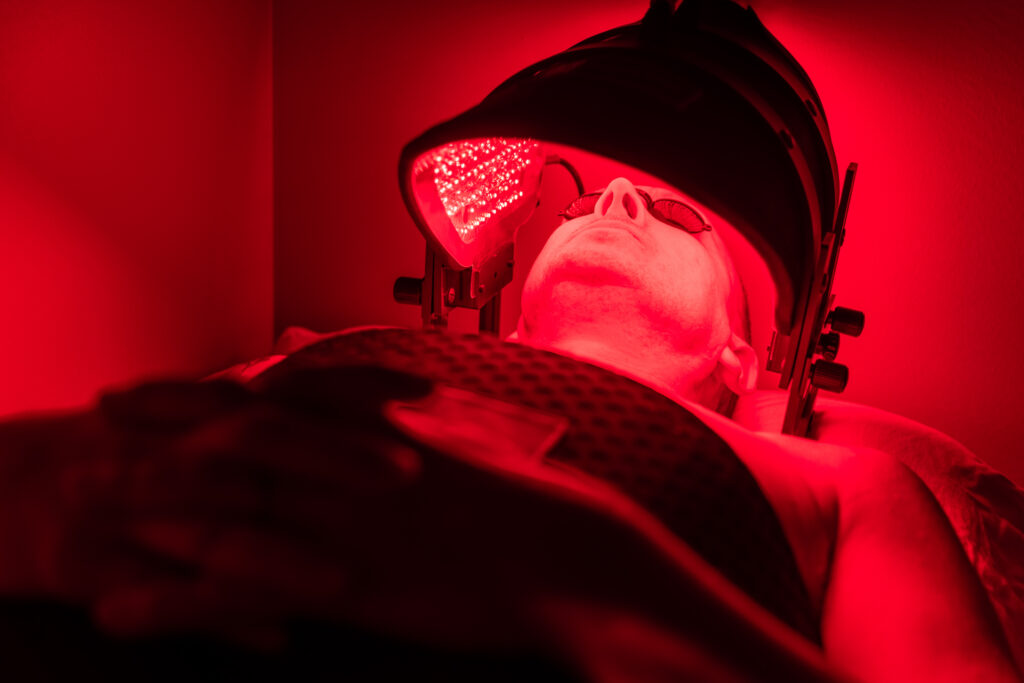 Patient lays on back with a laser cap placed above their head for red light therapy. The cap is emitting wavelengths of light to stimulate cellular function to promote healing and reduce inflammation. The patient is receiving this service after asking their local chiropractor about how to reduce inflammation in the body.