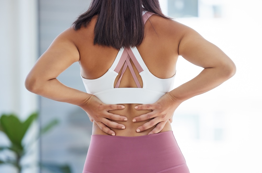 Woman in yoga gear places both hands on lower back to pinpoint the source of pain in lower back.