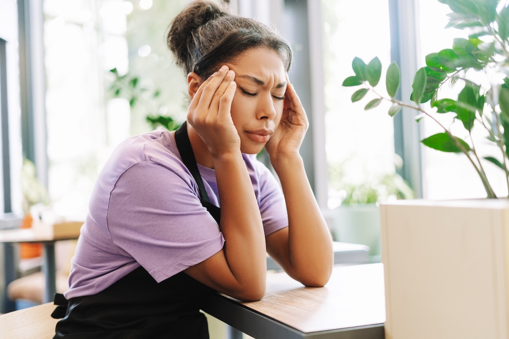 Woman sits at a desk with hands on her temple, struggling with headaches and migraines, which can be treated with neck pain relief near Iowa City